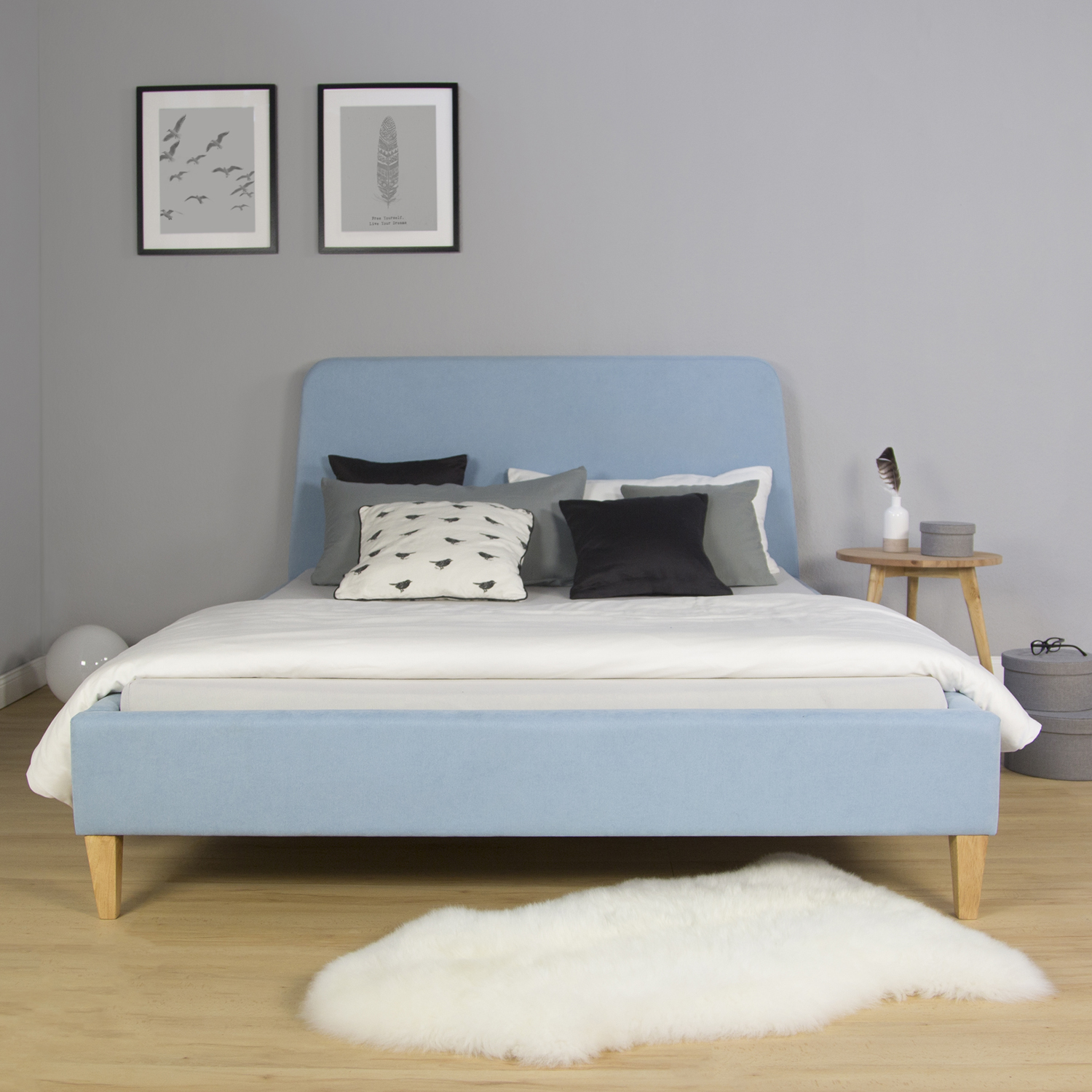 Polsterbett in Blau mit Stoffbezug. Das hellblaue Polsterbett mit abgerundetem Kopfteil und naturfarbenen Füßen ist mit Matratze, heller Bettwäsche, dunklen Kissen und einer grauen Decke vor weißem Hintergrund abgebildet.