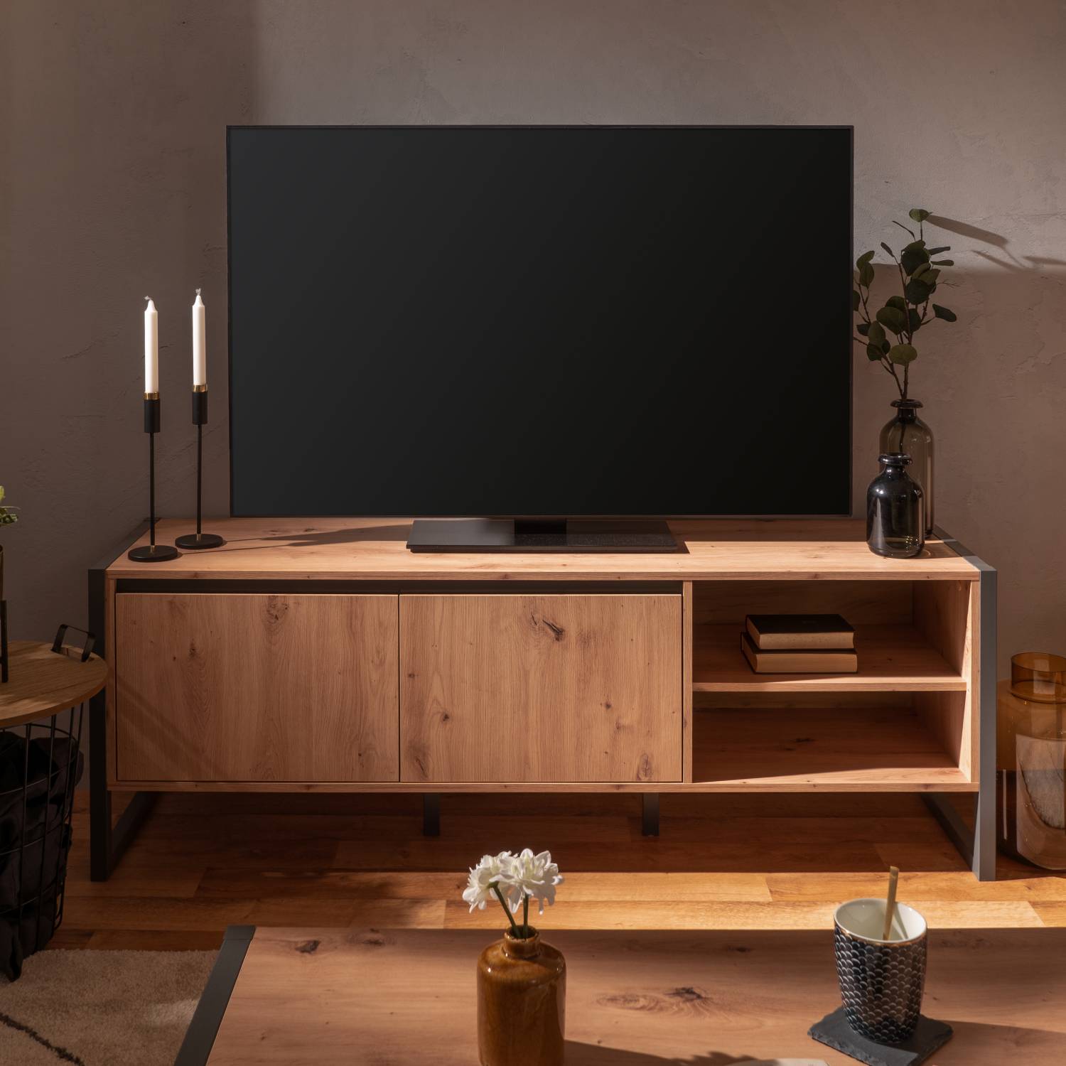 TV-Lowboard in Grau und Naturholzfarbe mit Holzelementen. Zwei geschlossene Türen links und zwei offene Fächer rechts sind vor weißem Hintergrund sichtbar. Das schwarze Metallgestell rahmt den niedrigen rechteckigen Holzkorpus ein; die Oberseite bleibt oh