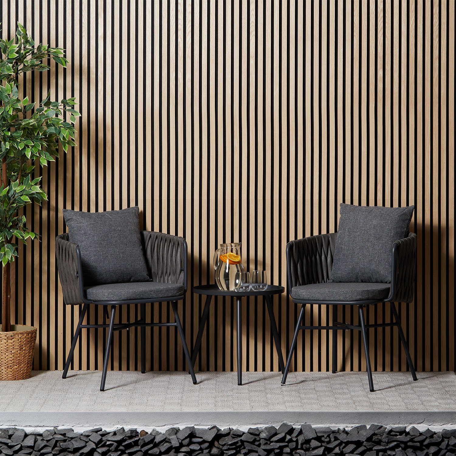 Gartenmöbel-Set mit Tisch und zwei Stühlen in Grau mit runder Tischform. Zwei dunkle geflochtene Sessel mit grauen Sitz- und Rückenkissen und ein runder schwarzer Beistelltisch sind zusammen auf weißem Hintergrund abgebildet. Armlehnen, schlanke Beine und
