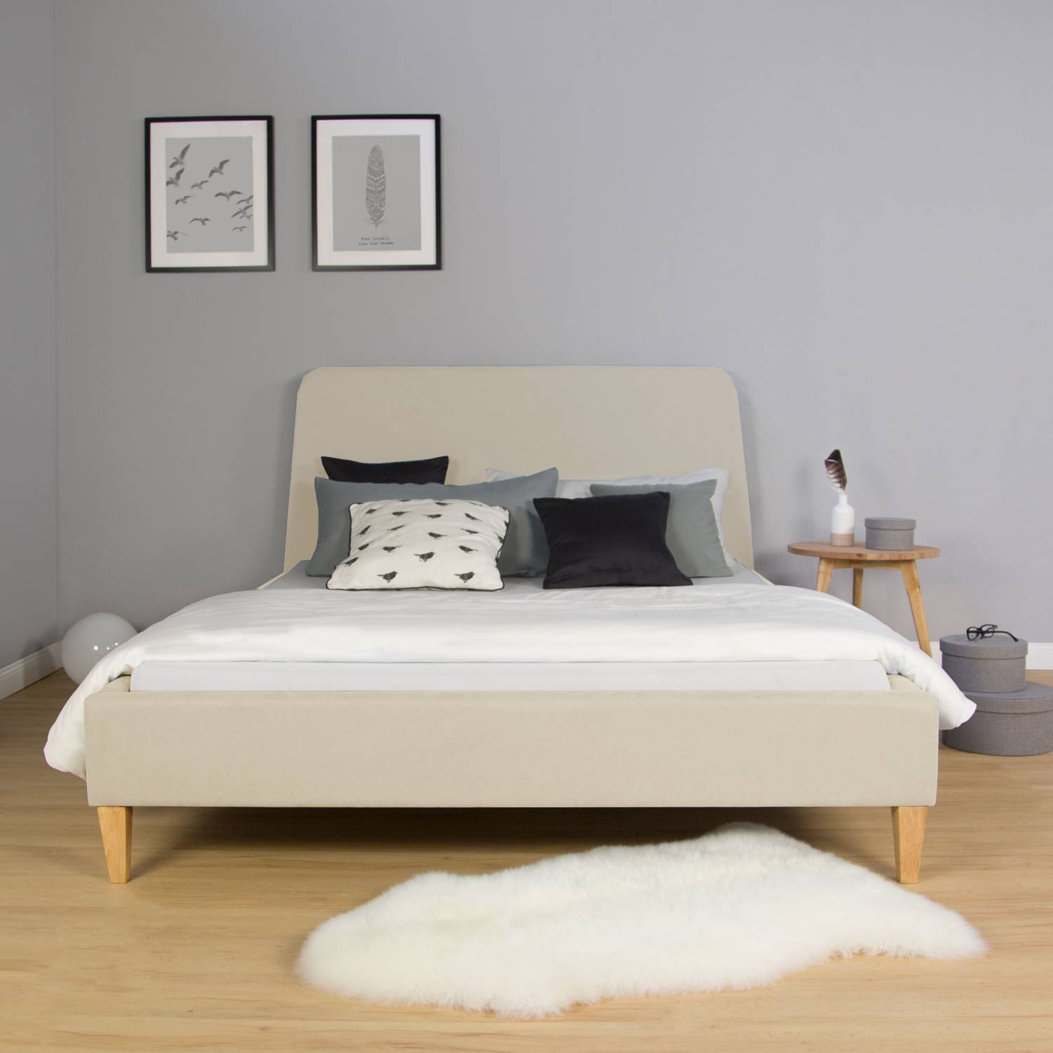 Polsterbett in Beige mit Stoffbezug. Das beigefarbene Polsterbett mit abgerundetem Kopfteil und naturfarbenen Füßen ist mit Matratze, heller Bettwäsche, dunklen Kissen und einer grauen Decke vor weißem Hintergrund abgebildet.