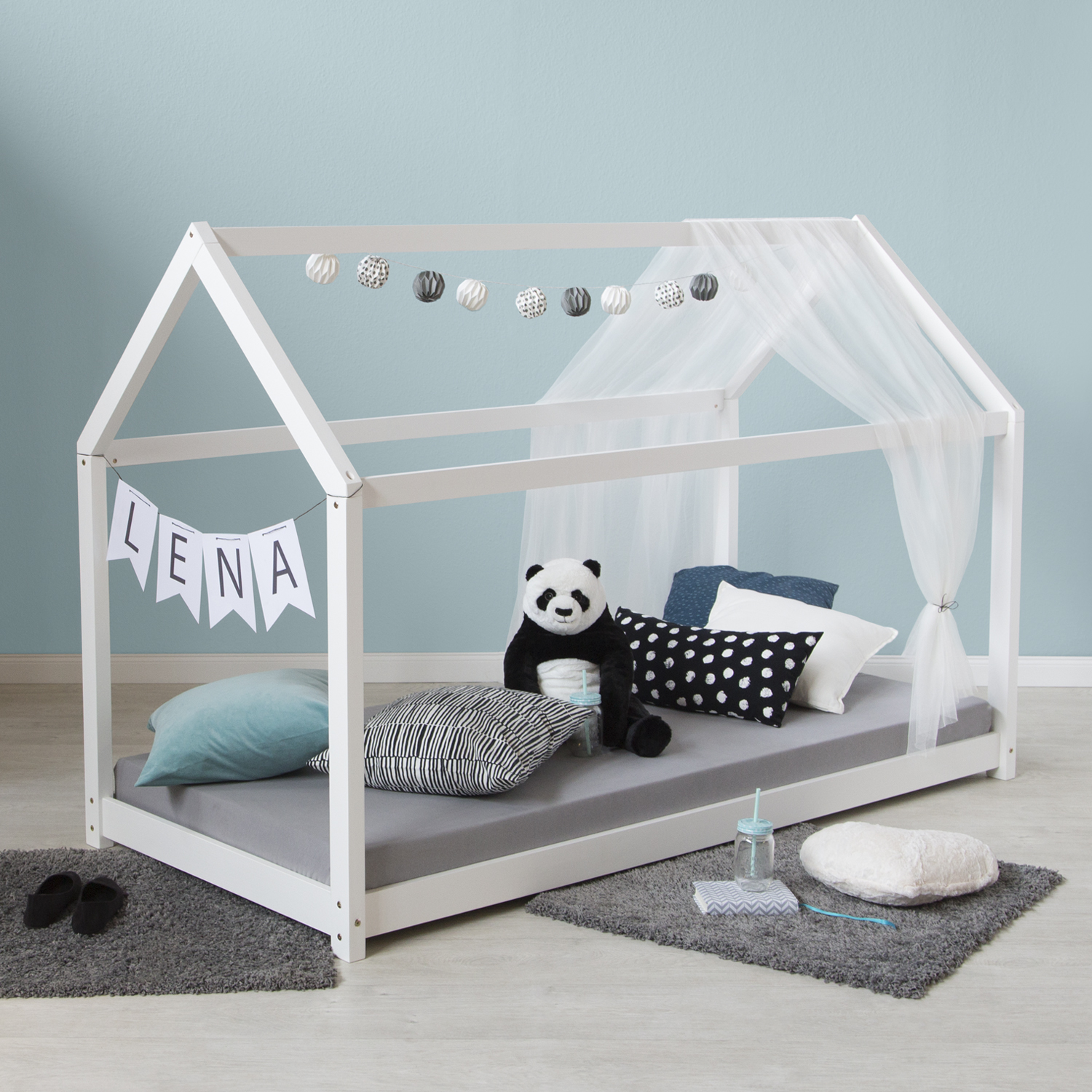 Kinderbett mit hausförmigem Rahmen in Weiß mit Holzelementen. Das Hausbett mit offenem Dachrahmen und niedrigen Seitenteilen ist mit Matratze und Kissen auf weißem Hintergrund abgebildet. Die Dachform und die Liegefläche sind in der schrägen Ansicht sicht