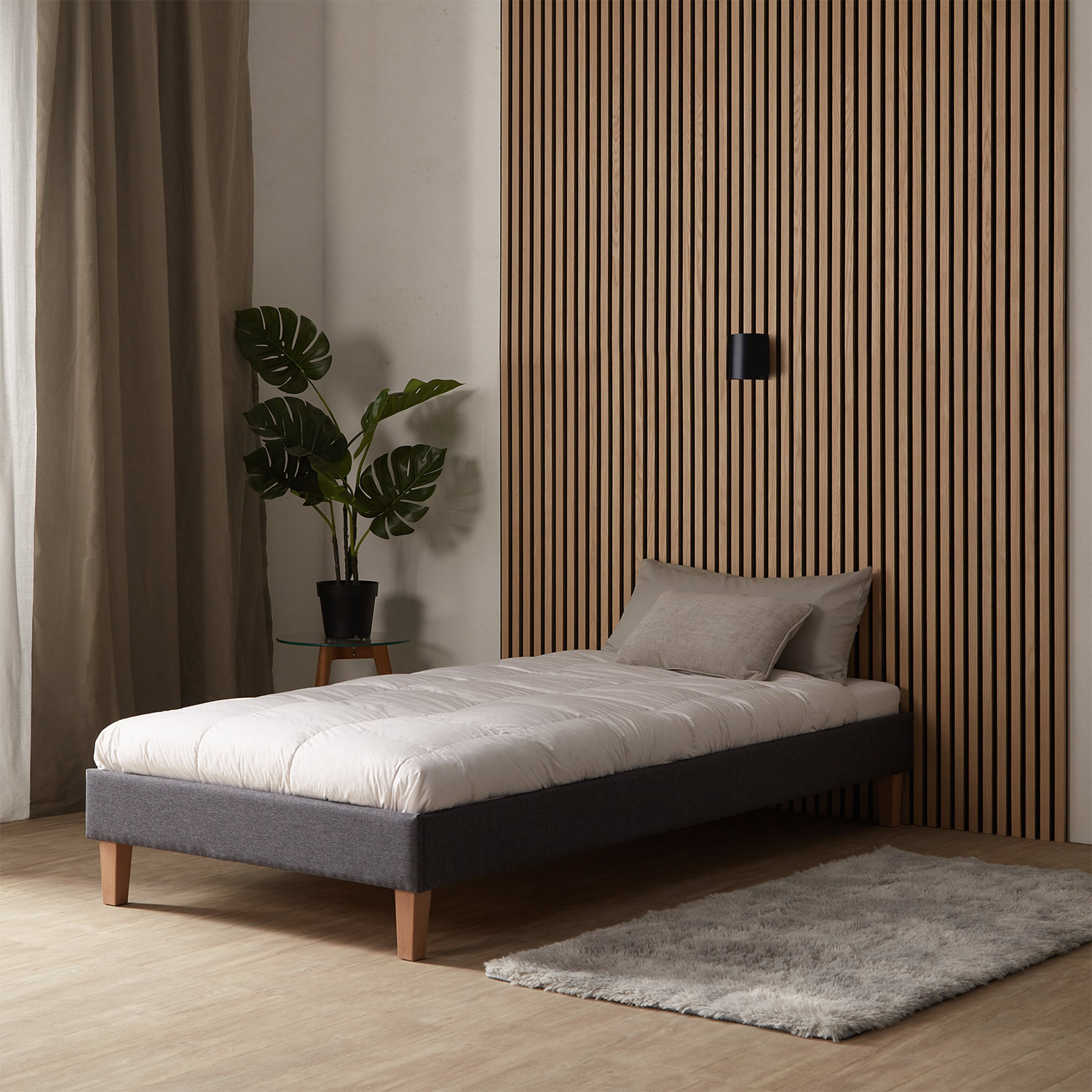 Polsterbett in Grau mit Stoffbezug, Lattenrost und Matratze. Das Bett ist mit Matratze, Bettwäsche und Kissen vor weißem Hintergrund abgebildet. Kopfteil, Rahmen und Beine sind in der schrägen Ansicht sichtbar.