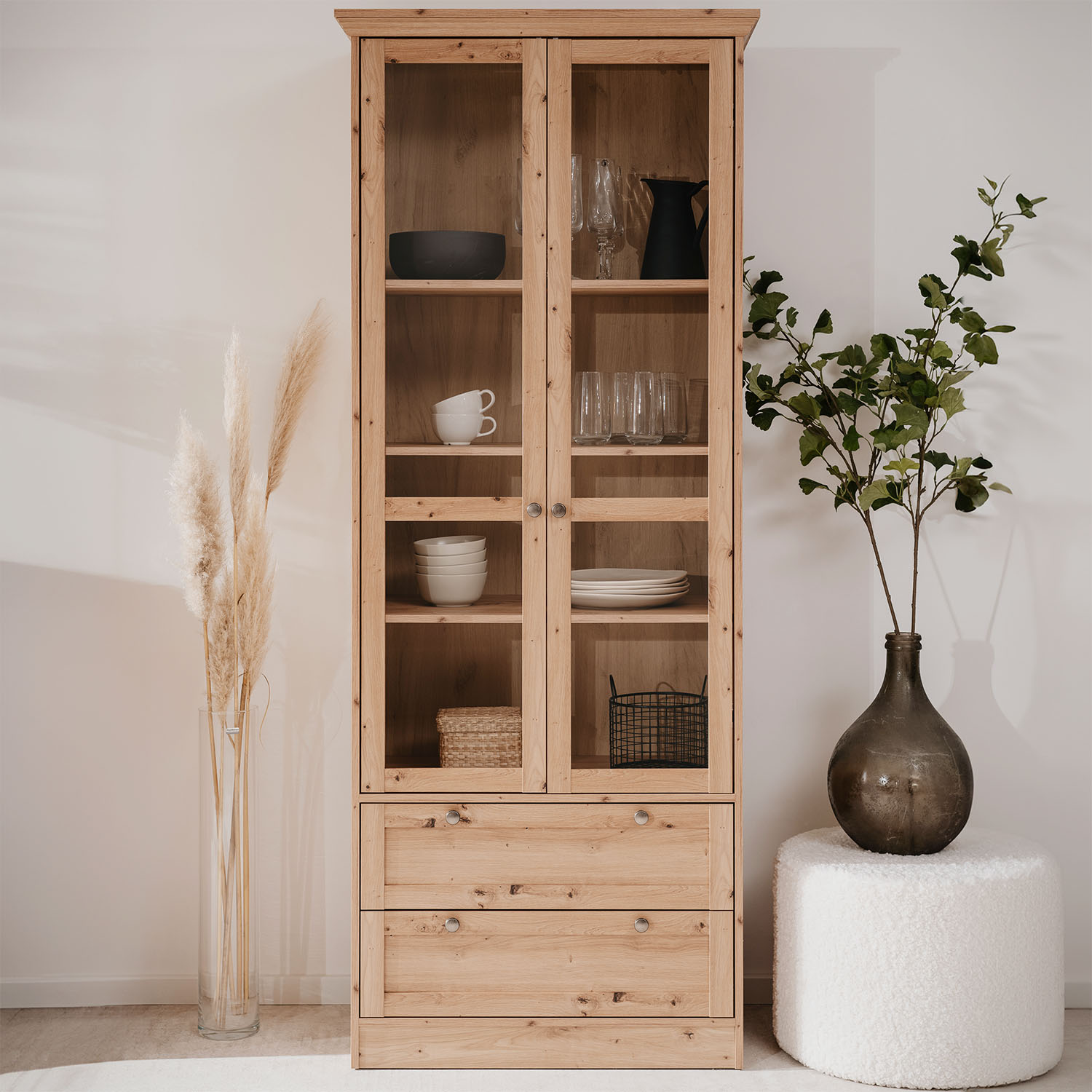Vitrine mit Oberflächen in Eichenoptik, Schubladenstauraum, Gestaltung im Landhausstil und Glaselementen. Der Artikel ist freigestellt auf weißem Hintergrund abgebildet. Sichtbare Details sind geschlossene Schranktüren, Schubladenfronten und eine sichtbar