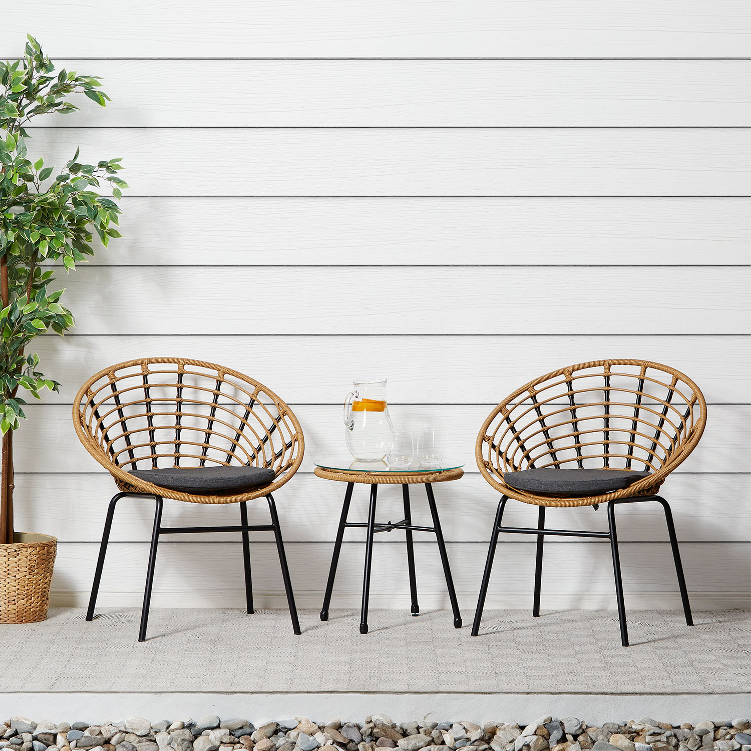 Gartenmöbel-Set mit Tisch und zwei Stühlen in Naturholzfarbe mit runder Tischform. Zwei naturfarbene Sessel mit offenem Flechtmuster, dunklen Sitzkissen und schwarzen Beinen stehen mit einem kleinen runden Beistelltisch vor weißem Hintergrund.