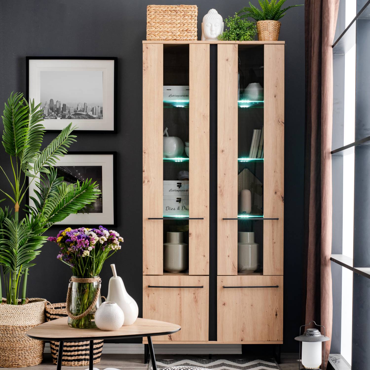 Vitrine in Schwarz und Naturholzfarbe mit Oberflächen in Eichenoptik. Der Artikel ist freigestellt auf weißem Hintergrund abgebildet. Sichtbare Details sind geschlossene Schranktüren, offene Ablagen, ein schwarzes Metallgestell und eine sichtbare Holzmase